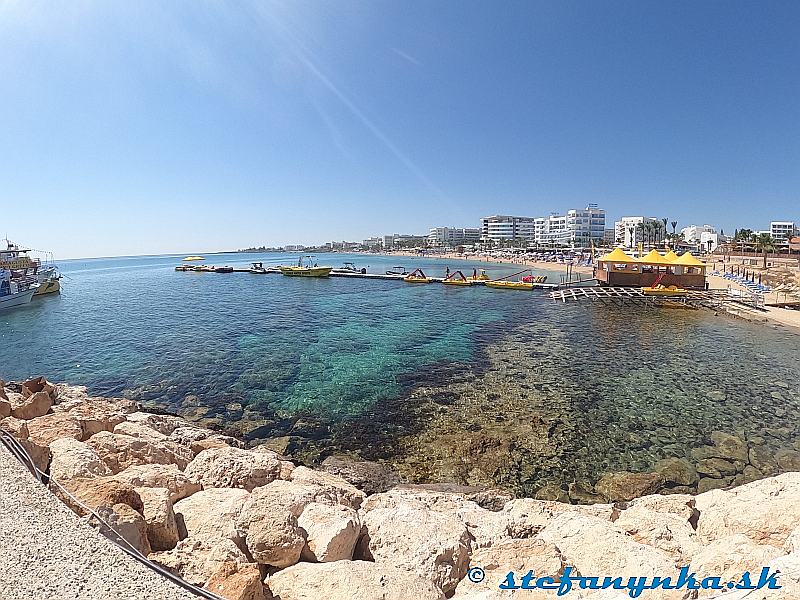 Protaras Pier - pohľad na pláž v Protarase. Hotel Silver Sands je takmer na konci (skoro nad špicou toho žltého motorového člna v stredisku vodných radostí na severnom konci pláže v Protarase). Ten posledný výbežok pevniny na obzore je už Fig Tree bay