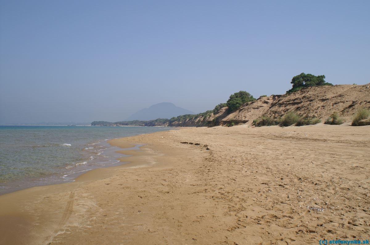 Agios Georgios south, Korfu. Záver pieskovej pláže. Pred výbežkom zelene do mora na ľavej strane sa nachádza vtok do jazera Korison. V pozadí kopec Pantokrator (ten južný)